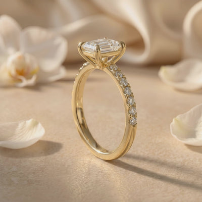 Side view of a gold-tone ring on a surface, showing the raised emerald-cut setting and pavé stones along the band.