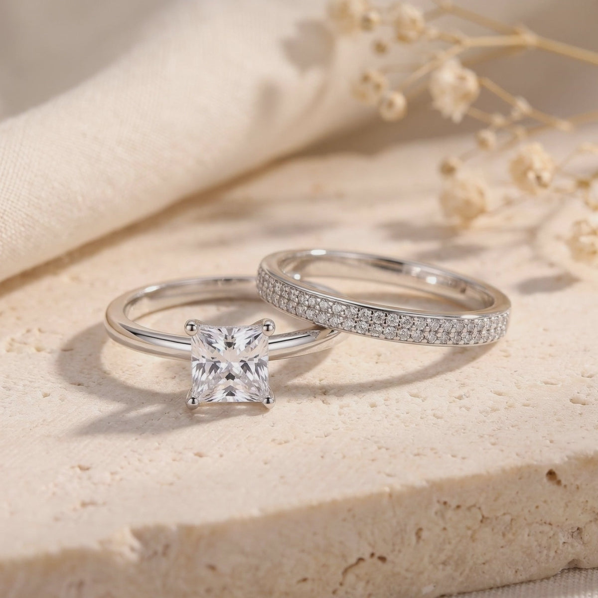 Silver-tone princess-cut solitaire and pavé band displayed together on a textured beige stone surface.
