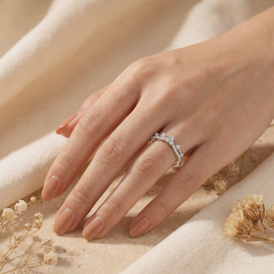 Tiara-style ring with clear stones shown worn on a hand over beige fabric, with dried flowers nearby.