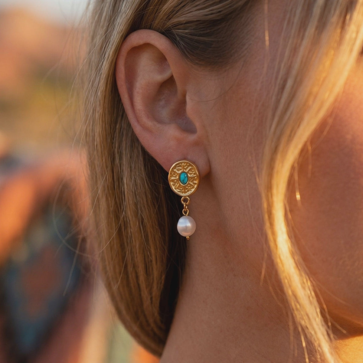 Close-up of a person wearing a gold-tone sunburst earring with a turquoise-hued center stone and simulated pearl drop.