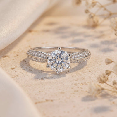 Close-up of a six-prong round clear stone ring resting upright on a textured beige surface.