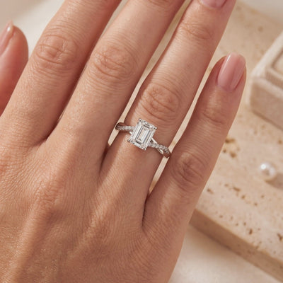 Emerald-cut silver solitaire ring with a twisted pavé band worn on a hand.