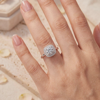 Silver ring featuring a round center stone and double cushion halo, worn on a hand.