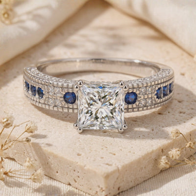 Front close-up of a silver ring with a princess-cut clear center and blue accent stones on a porous beige slab.