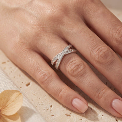 Silver crossover ring with clear pavé stones worn on a finger over a textured stone surface.
