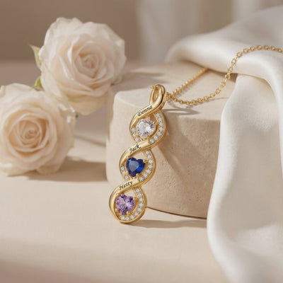 Gold-toned vertical infinity necklace with three heart birthstones and engraved names, displayed beside white roses.
