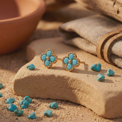 Pair of gold-toned four-petal turquoise stud earrings displayed on a stone over sand.