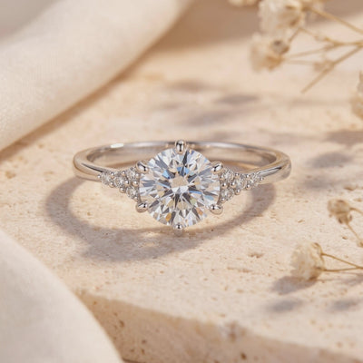 Silver ring with a round clear center stone and trefoil shoulder accents, set on a beige stone surface.