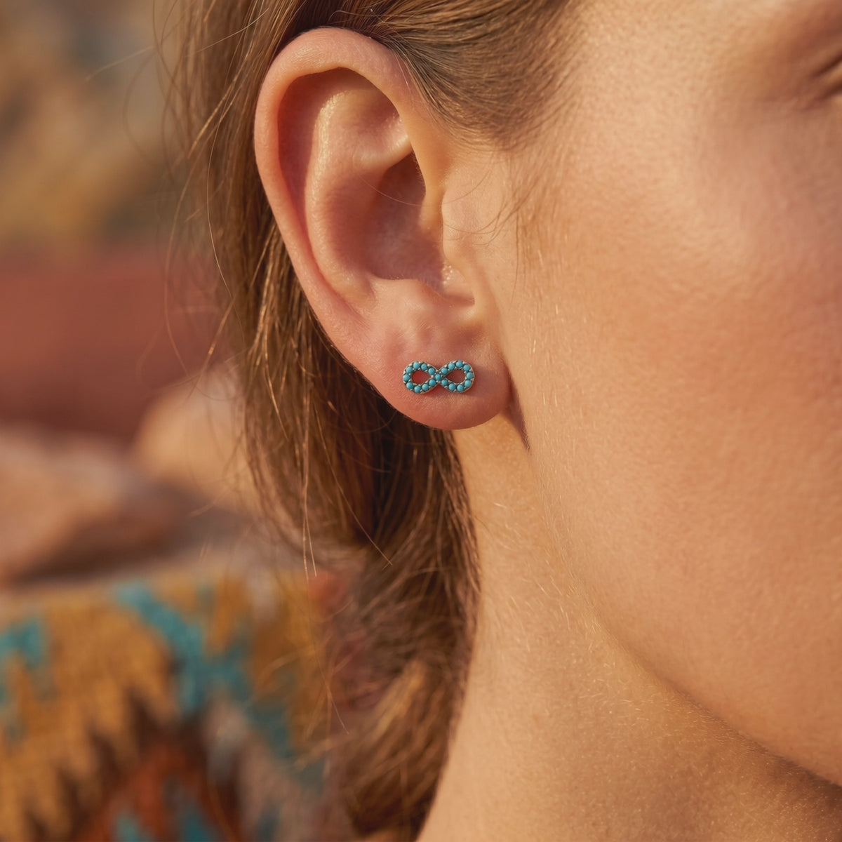 Close-up of an ear wearing a silver infinity stud earring with small turquoise stones.