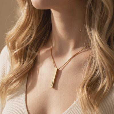 Gold-toned bar pendant necklace worn on a model’s neck, with dark engraved text on the pendant.