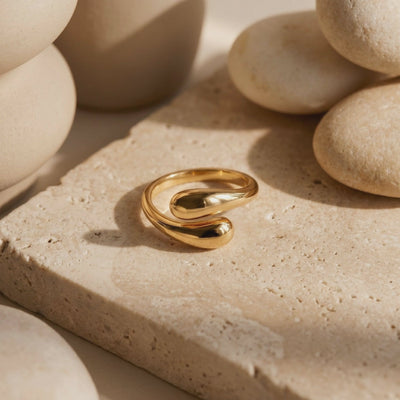 Gold-tone open ring with rounded ends resting on a textured beige stone slab among smooth stones.