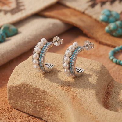 Pair of silver C-hoop earrings with pearls, blue turquoise, and clear accents displayed on a textured stone block.