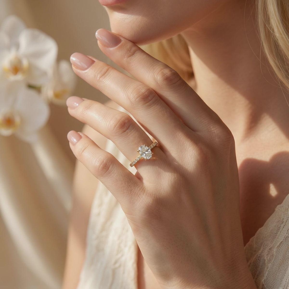 Oval-cut solitaire ring with a pavé yellow gold-tone band worn on a hand beside white orchids.