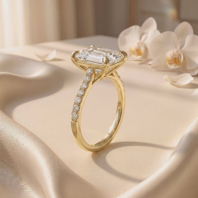 Yellow-toned emerald-cut pavé solitaire ring shown in side view on satin fabric, with orchids in the background.