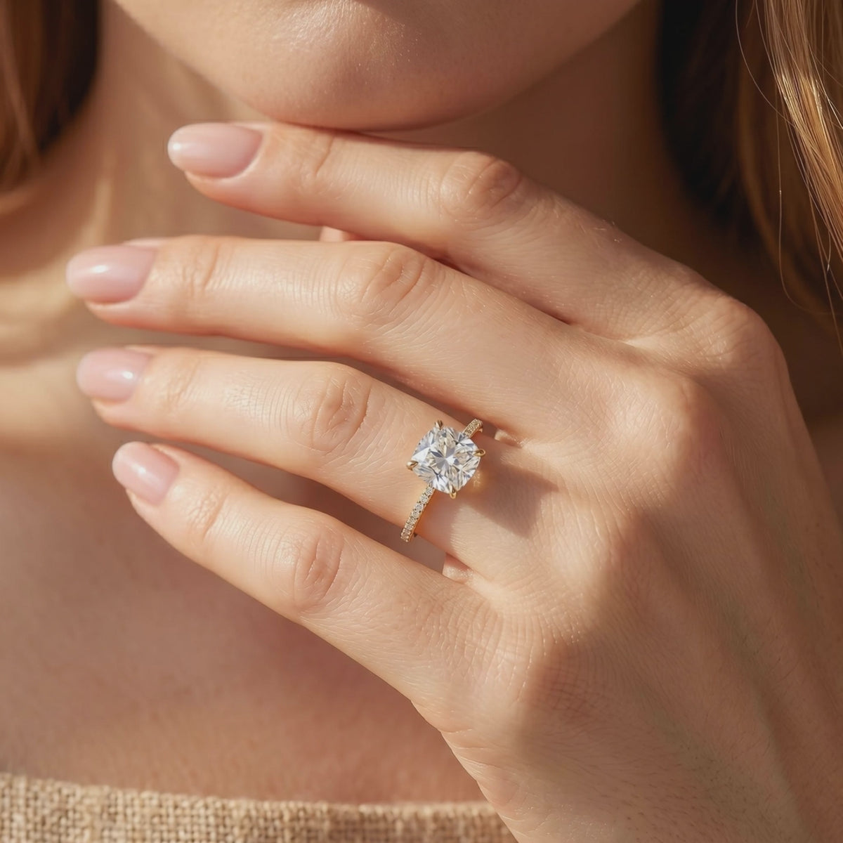 Yellow gold-tone cushion-cut solitaire ring with a pavé band worn on a hand near the wearer’s face.