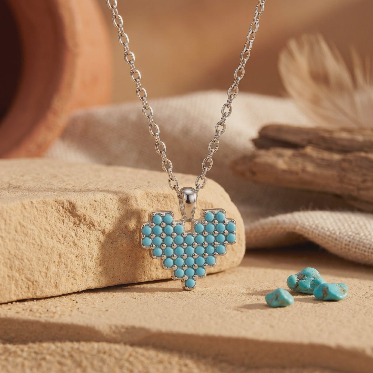 Close-up of the silver necklace and turquoise heart pendant displayed on a beige stone surface beside loose turquoise pieces.