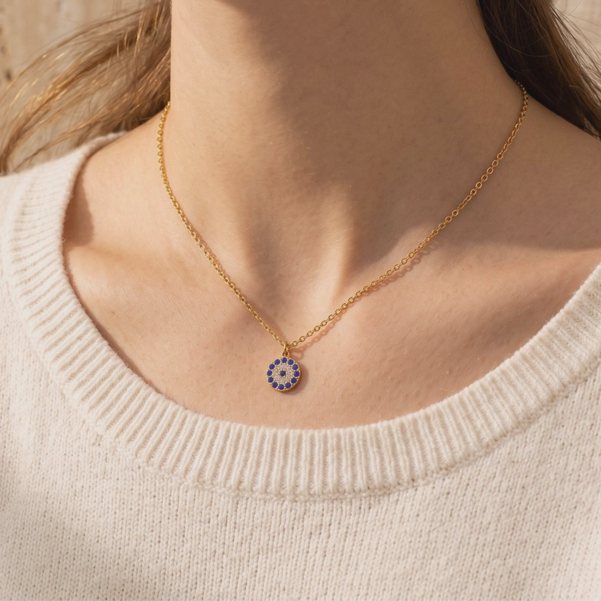 Gold-toned chain necklace worn at the neck, with a round blue-and-white pavé pendant over a cream ribbed sweater.