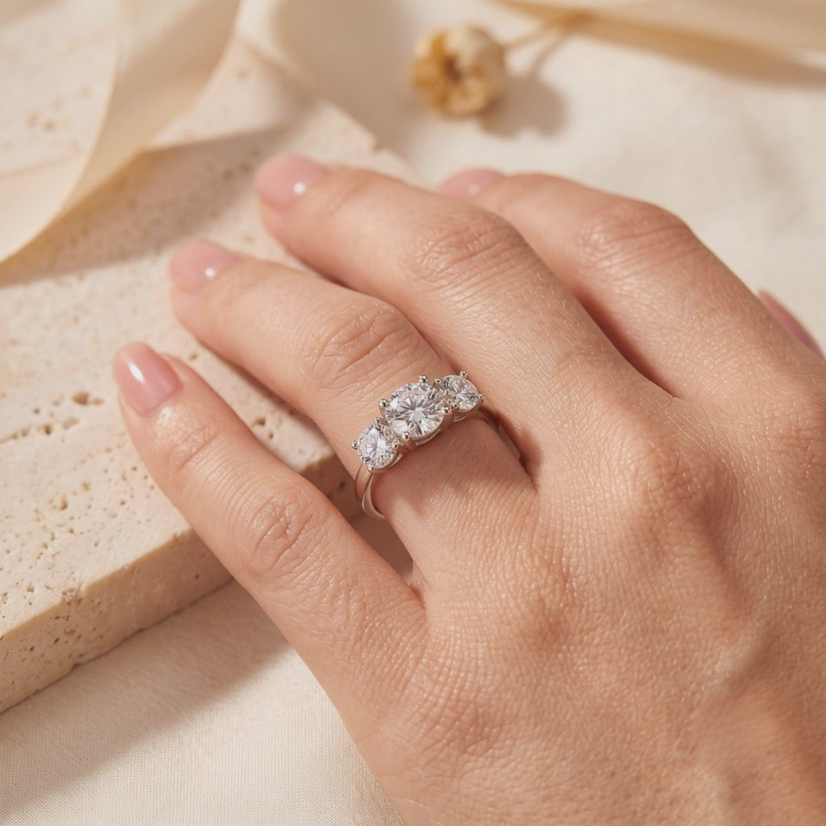 Silver three-stone ring with clear round stones worn on a hand over a beige stone slab and fabric.