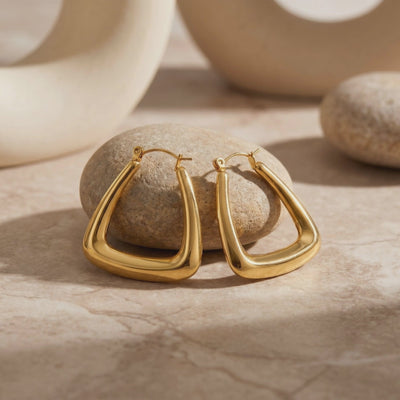 Pair of gold-toned triangular hoop earrings displayed in front of a rounded stone on a pale tabletop.