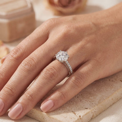 Silver solitaire ring with a round clear stone in a six-prong setting, worn on a finger against a warm background.
