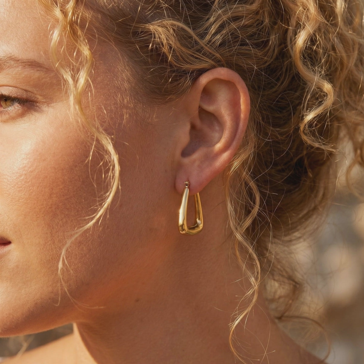 Side profile of a person with curly hair wearing a chunky gold-toned triangular hoop earring.