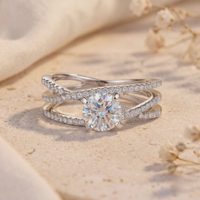 Close-up of a silver criss-cross ring resting on a textured surface, with a round center stone and pavé bands.