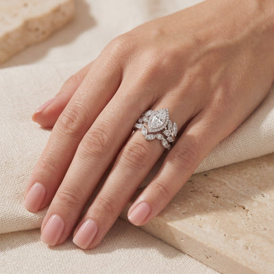 Silver marquise-cut halo ring stacked with a leaf-motif band on a hand against beige fabric.