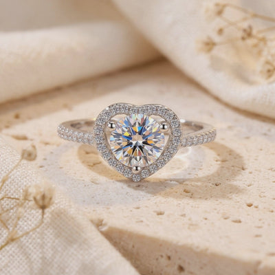 Close-up of a silver ring with a round center stone inside a heart-shaped halo on a textured stone surface.