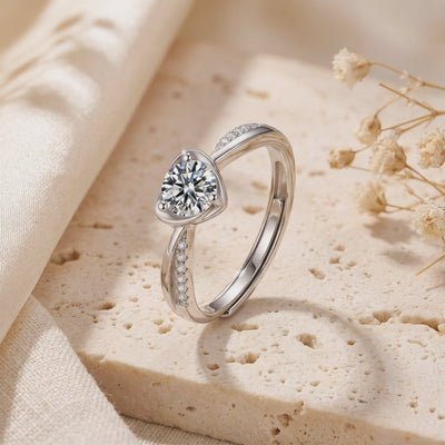 Silver heart-bypass ring with a round center gem and pave accents, standing on a textured stone surface.