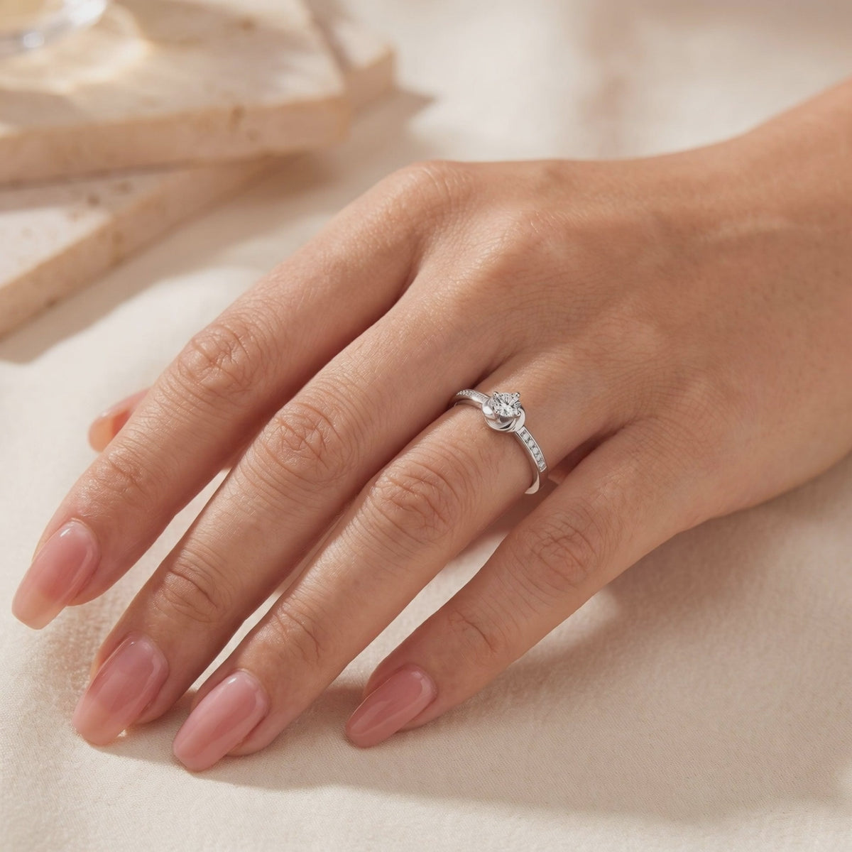Silver Melisande bypass ring with a round clear center stone and accent stones, worn on a hand against beige fabric.