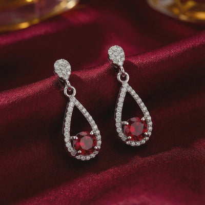 Pair of solid white gold teardrop earrings with round red simulated ruby center stones and clear pavé halos on deep red satin.