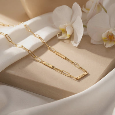 Gold link chain necklace with engraved name tags displayed on a stone slab beside white orchids.