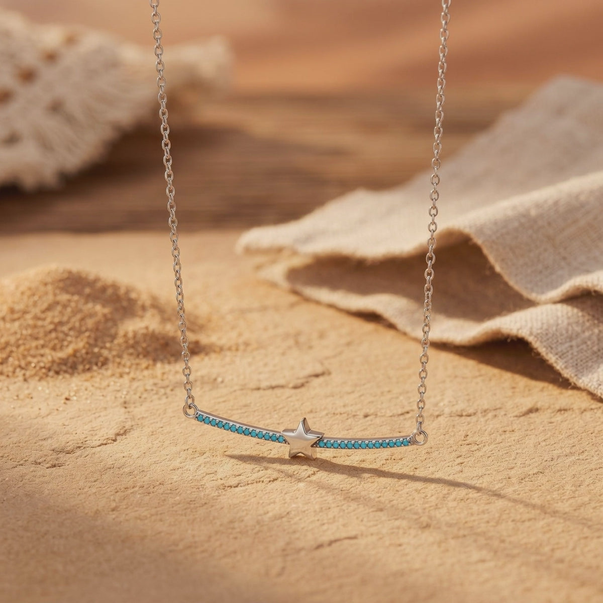 Silver necklace with a turquoise-set curved bar and center star, displayed on a tan textured surface beside beige fabric.
