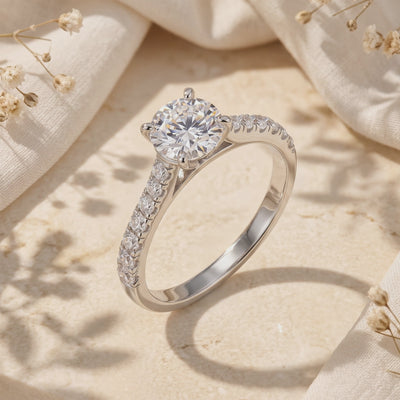 Silver solitaire ring with a round clear center stone and pavé shoulders standing on a beige textured surface.
