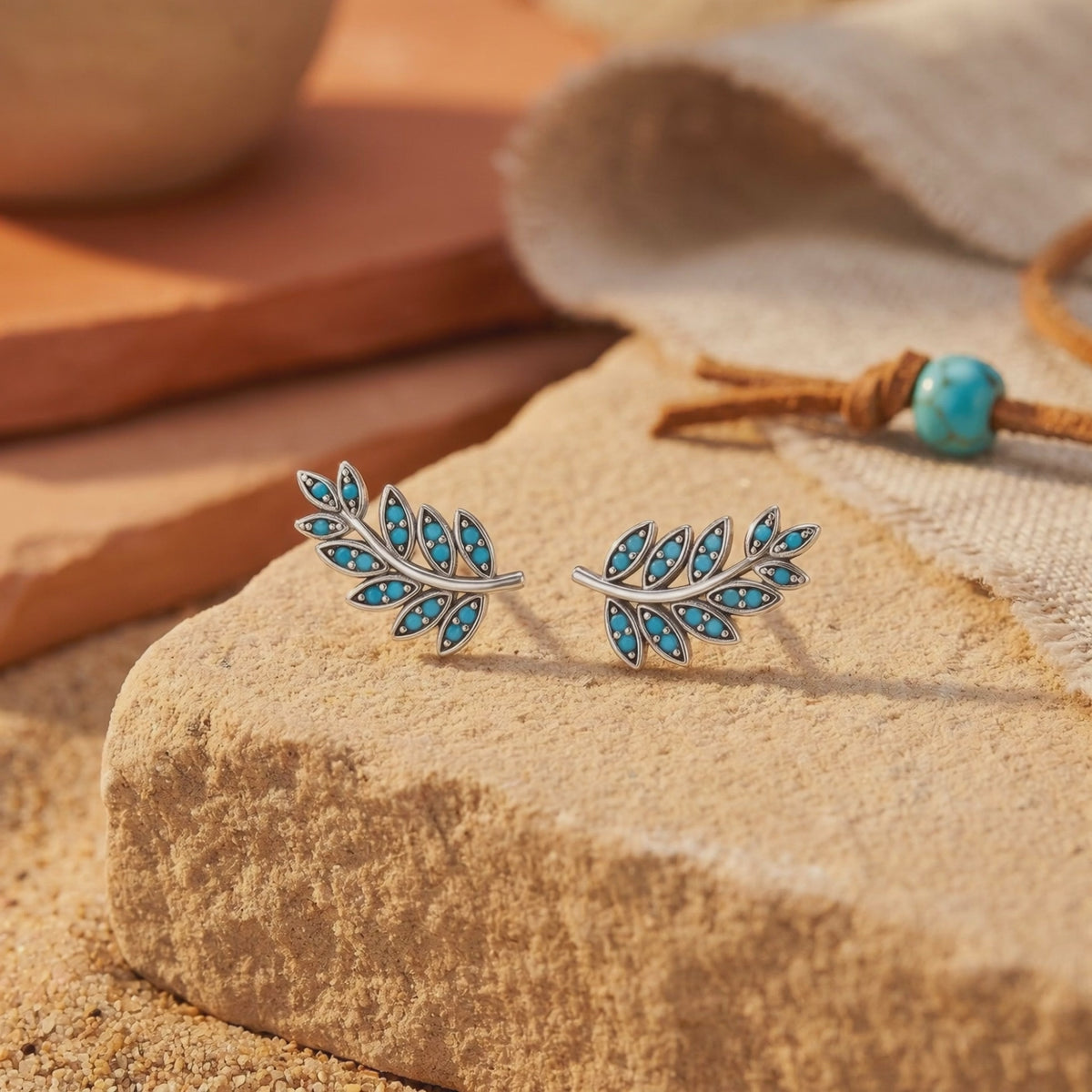 Pair of silver leaf-branch earrings with turquoise stones displayed upright on a tan stone block.