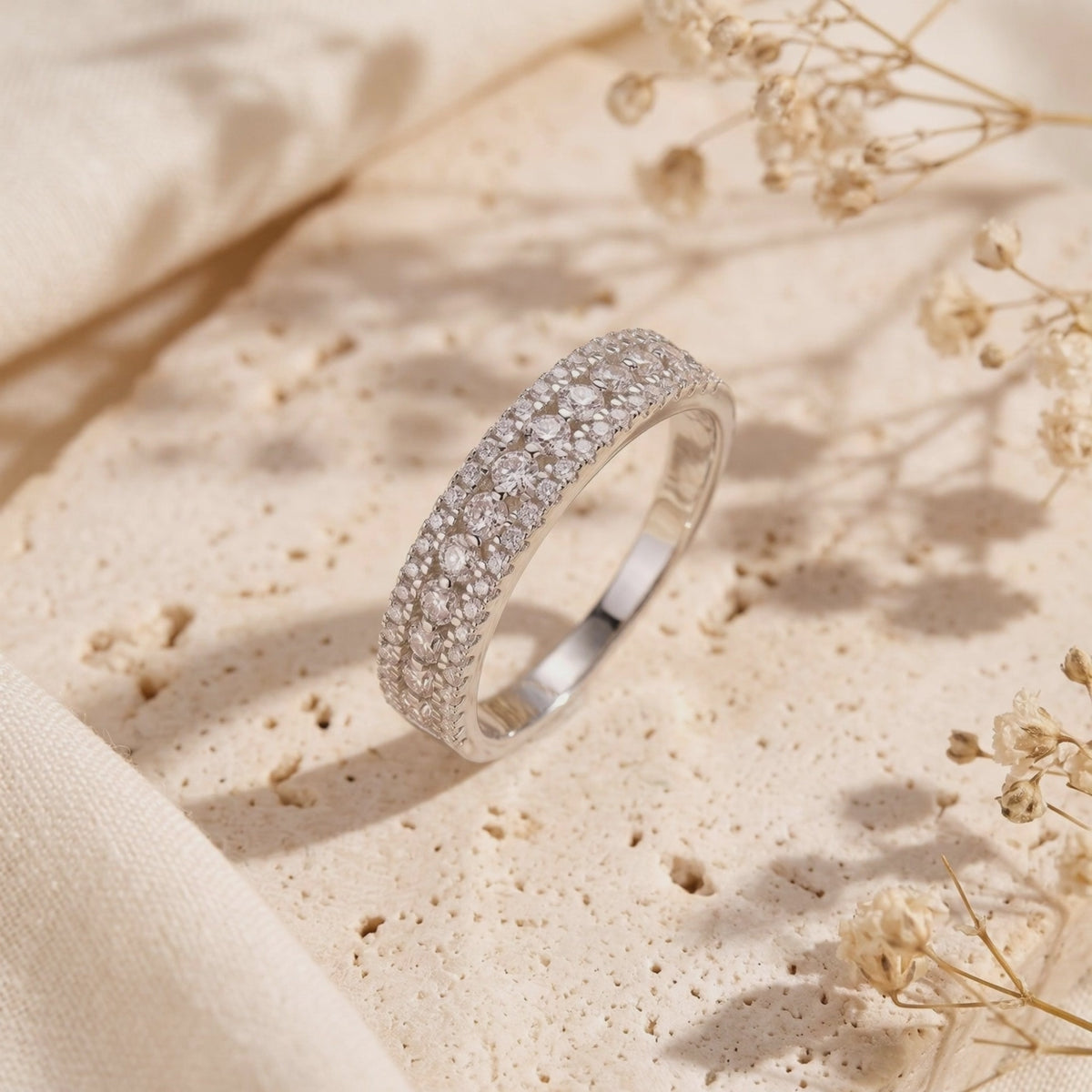 Three-row silver pavé ring propped upright on a textured stone surface with dried flowers nearby.
