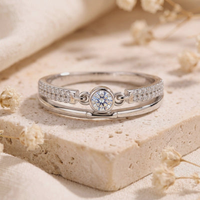 Silver double-band ring displayed upright on a beige stone block, showing its round center stone and polished lower band.