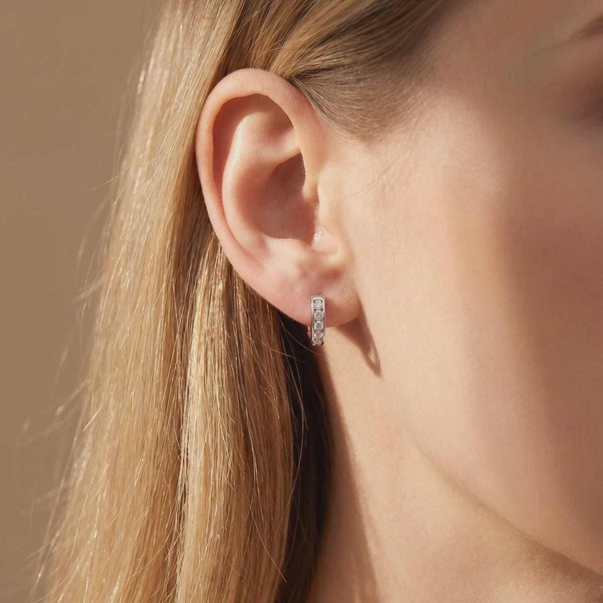 Silver huggie hoop earring with channel-set round clear stones worn on a blonde model's earlobe in profile.