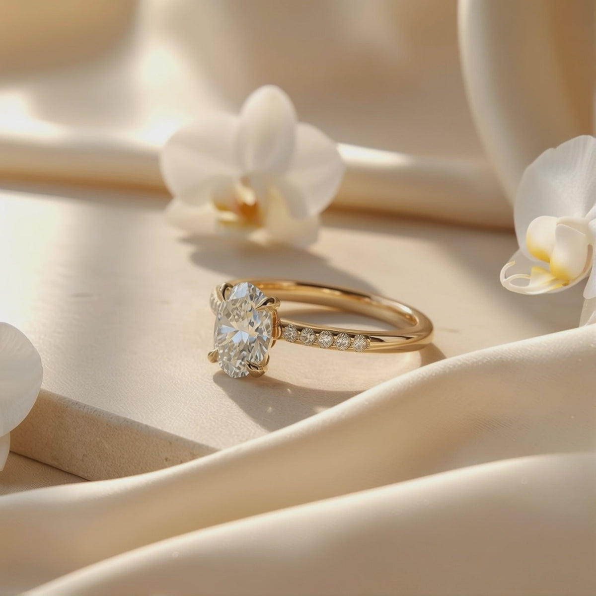 Yellow gold-tone oval solitaire ring with a pavé band resting on beige satin among white orchids.