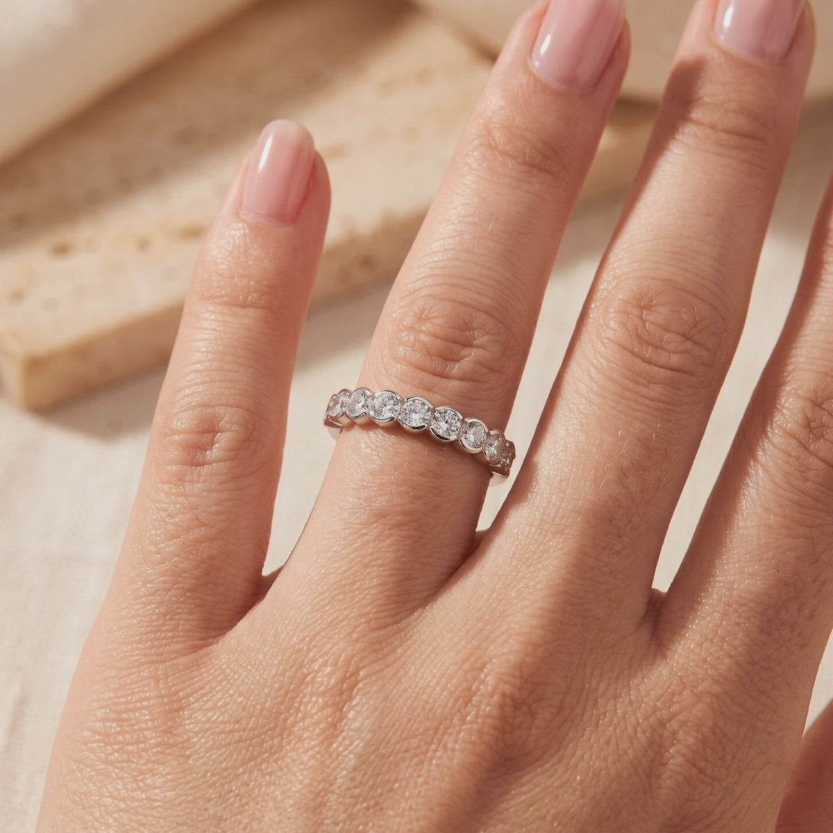 Silver bezel-set round clear-stone band worn on a hand against a warm beige background.