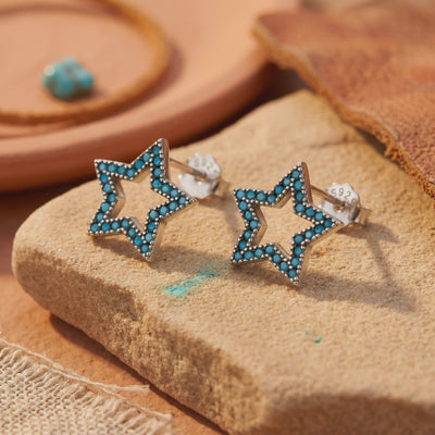 Pair of silver open star stud earrings with turquoise pavé stones displayed on a textured tan stone surface.