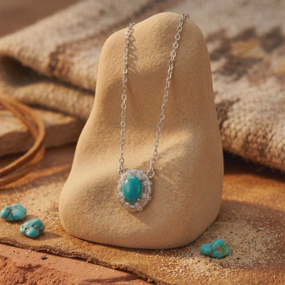 Silver turquoise pendant necklace displayed on a beige stone amid loose turquoise stones in a desert-inspired setting.