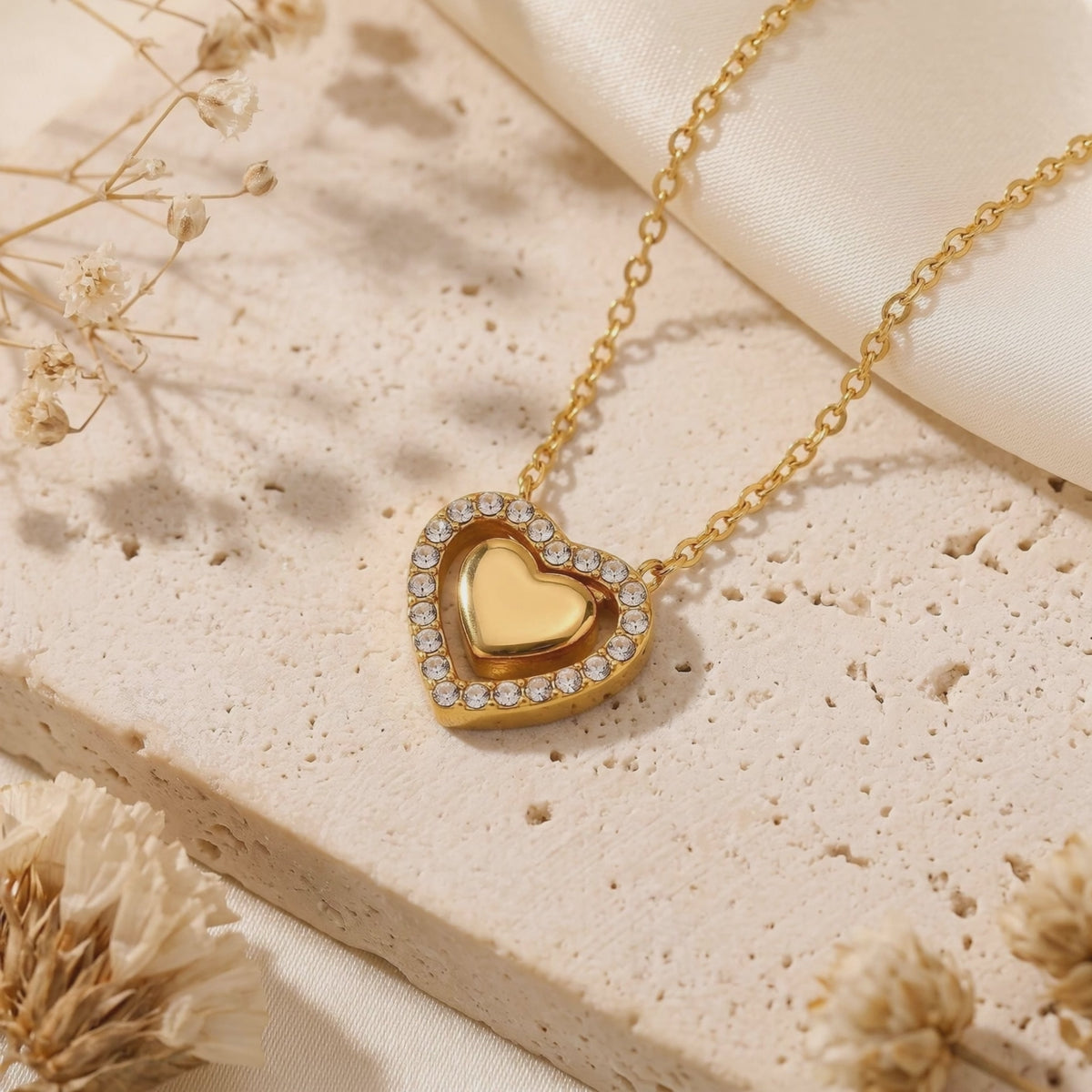 Close-up of the gold Priscilla double-heart pendant with cubic zirconia on a porous stone slab with dried flowers.