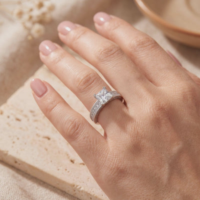 Silver ring with a princess-cut center stone and pavé band worn on a hand over a textured surface.