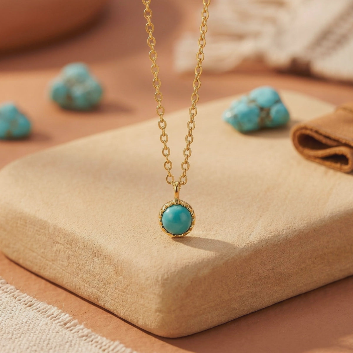 Close-up of a gold chain necklace with a round turquoise cabochon pendant displayed on a beige block.