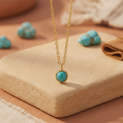 Close-up of a gold chain necklace with a round turquoise cabochon pendant displayed on a beige block.