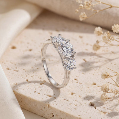 Angled view of a silver three-stone ring with clear round stones standing on a beige stone slab.