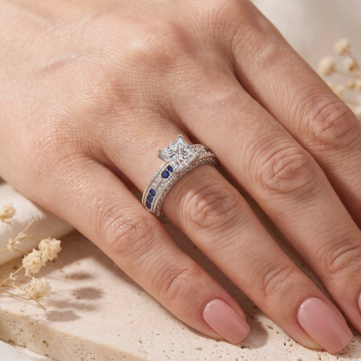 Princess-cut clear gem ring with blue sapphire-colored accents, worn on a hand near dried flowers.