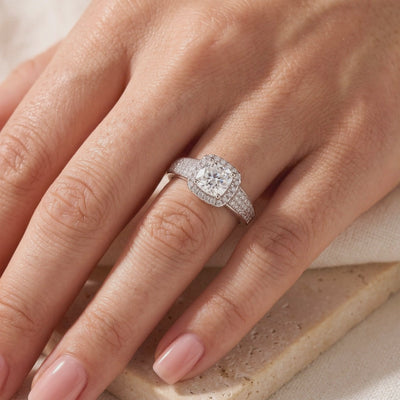 Silver cushion halo ring with a clear center stone and pavé band worn on a hand