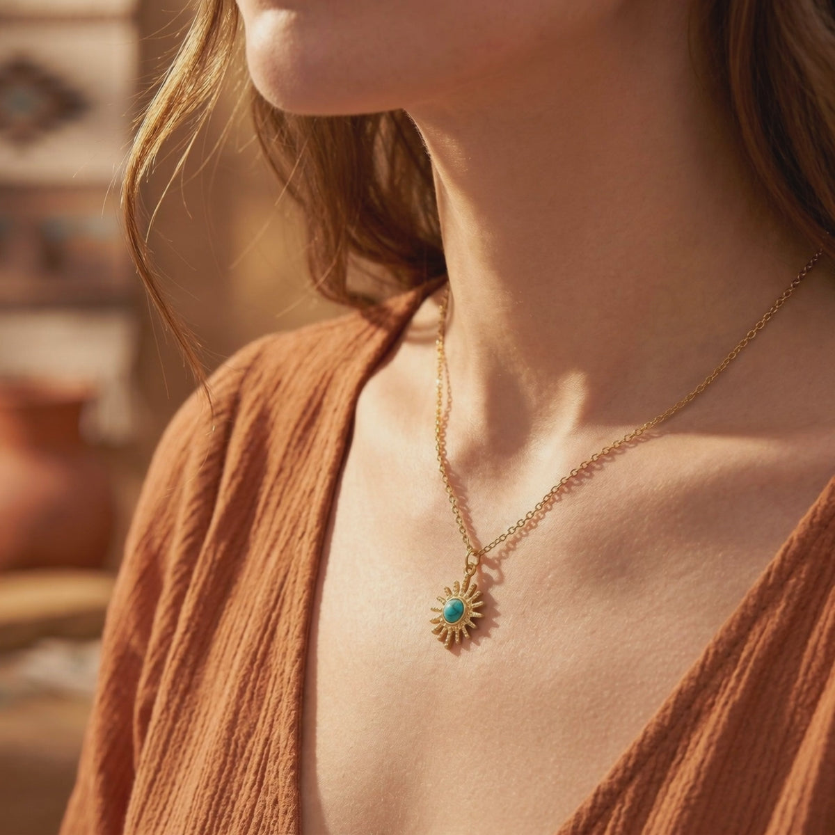 Model wearing a gold-toned sunburst pendant with a blue-green center cabochon against a rust-colored textured garment.